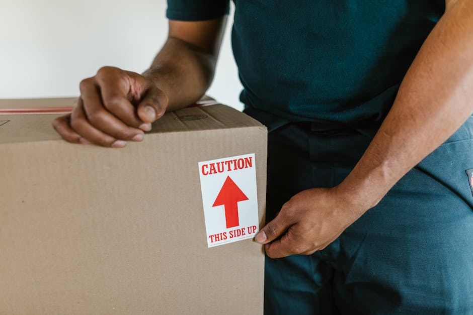 A man with curly dark hair and a beard, wearing a blue T-shirt and dark jeans, is seated on a wooden floor inside a room with white walls. He is leaning against a small stack of cardboard boxes that a